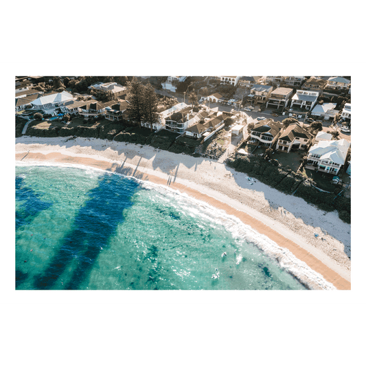 Aerial view of Blue Bay, NSW, featuring turquoise waters, golden sand, and beachfront homes with long afternoon shadows cast by Norfolk pines.