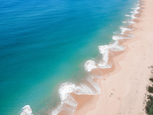 Aerial view of Copacabana Beach, NSW, showcasing rolling aqua waves curling onto golden sand with people scattered along the shoreline.
