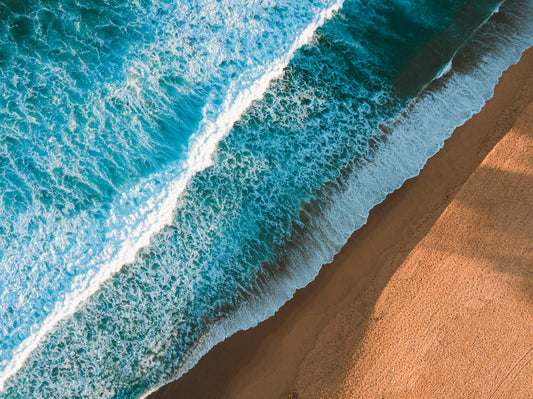 Aerial view of Copacabana Beach, NSW, showcasing deep blue waves crashing onto golden sand with long shadows creating stunning textures.
