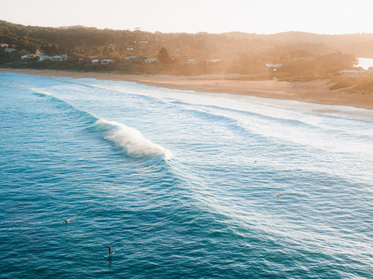 A golden sunset over Copacabana Beach, NSW, with rolling waves and surfers paddling through the deep blue water.
