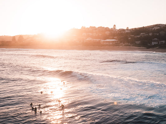 A golden sunset over Copacabana Beach, NSW, with surfers waiting in the waves as the sun reflects off the water.
