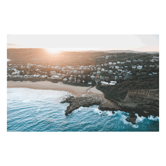 Aerial view of North Avoca Beach, NSW, at sunset, with golden light illuminating the coastal hillside, ocean waves, and rocky shoreline.