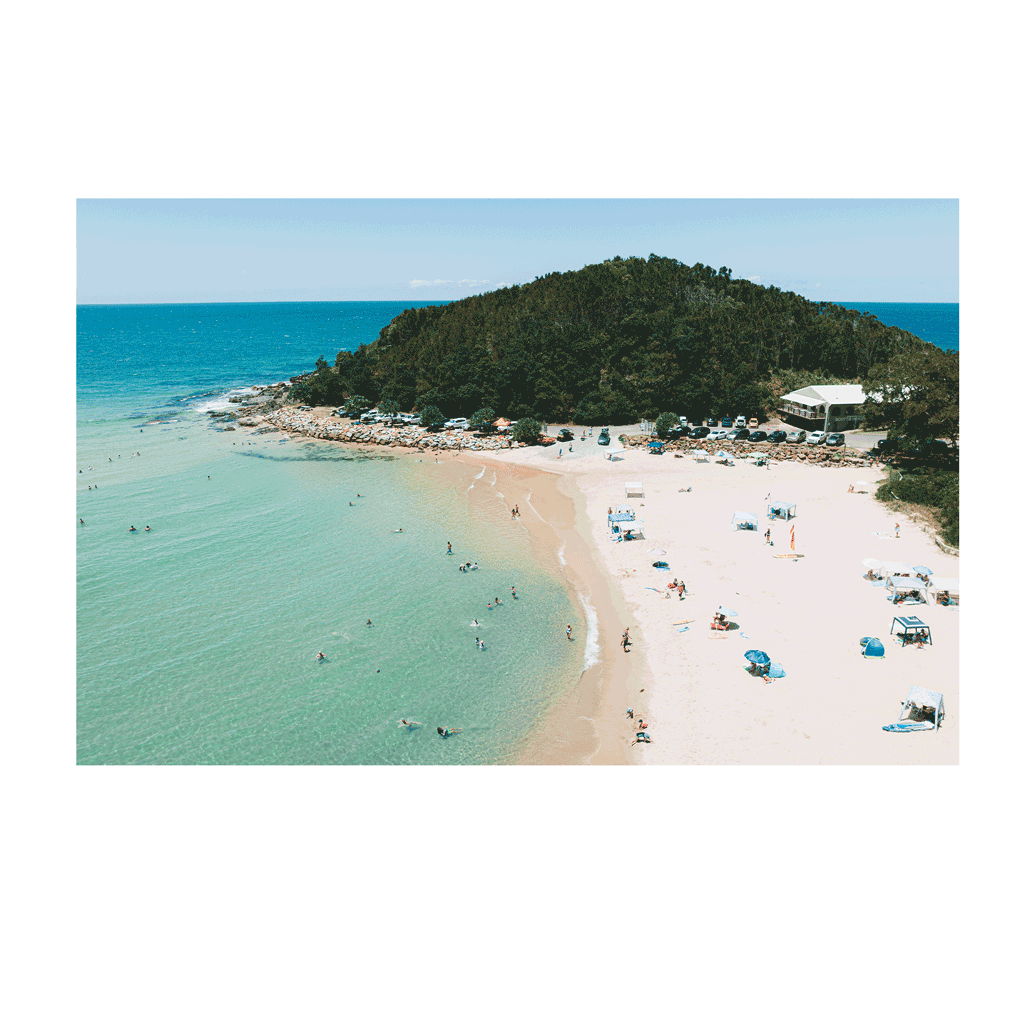 A picturesque view of Scotts Head, NSW, featuring golden sands, turquoise waters, and beachgoers enjoying a sunny day. The lush green headland and beachfront café complete this idyllic coastal scene.
