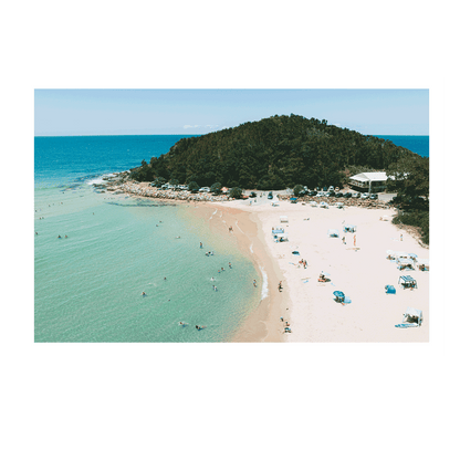 A picturesque view of Scotts Head, NSW, featuring golden sands, turquoise waters, and beachgoers enjoying a sunny day. The lush green headland and beachfront café complete this idyllic coastal scene.
