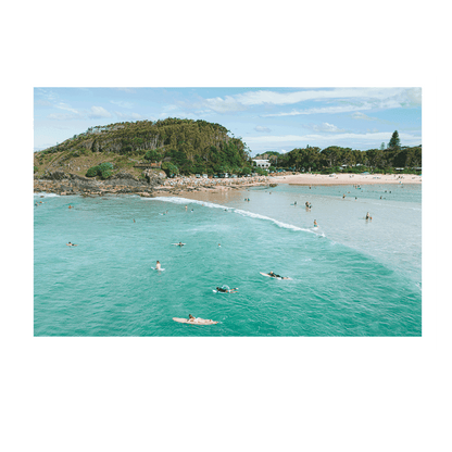 A dreamy, sunlit coastal scene at Scotts Head, NSW, featuring a surfer gliding across shimmering ocean waters. The blurred foreground of green coastal foliage adds depth to the tranquil seaside moment.
