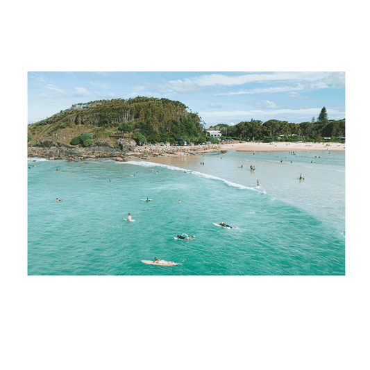 A dreamy, sunlit coastal scene at Scotts Head, NSW, featuring a surfer gliding across shimmering ocean waters. The blurred foreground of green coastal foliage adds depth to the tranquil seaside moment.
