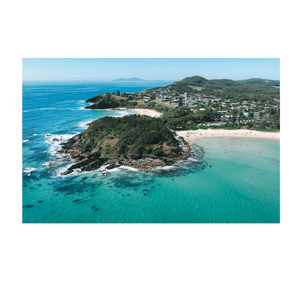 A breathtaking aerial view of Scotts Head, NSW, featuring turquoise waters, a rugged headland, and a sun-soaked beach with swimmers and surfers. Coastal homes and rolling green hills complete the picturesque landscape.
