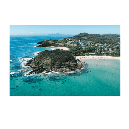 A breathtaking aerial view of Scotts Head, NSW, featuring turquoise waters, a rugged headland, and a sun-soaked beach with swimmers and surfers. Coastal homes and rolling green hills complete the picturesque landscape.
