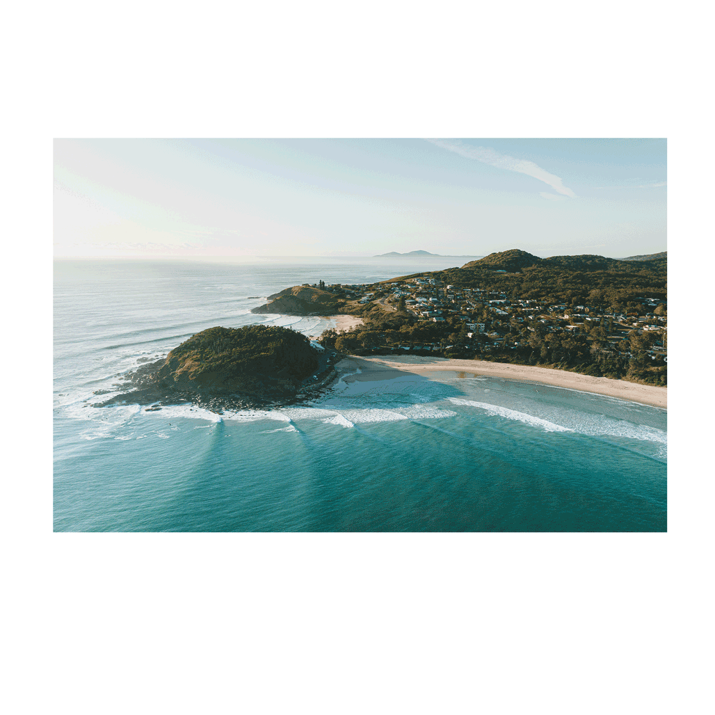 A breathtaking aerial view of Scotts Head, NSW, bathed in golden morning light. Waves roll onto the pristine beach as the coastal town nestles between lush green hills, creating a serene and picturesque seascape.
