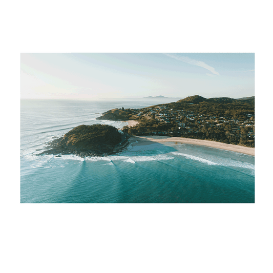 A breathtaking aerial view of Scotts Head, NSW, bathed in golden morning light. Waves roll onto the pristine beach as the coastal town nestles between lush green hills, creating a serene and picturesque seascape.
