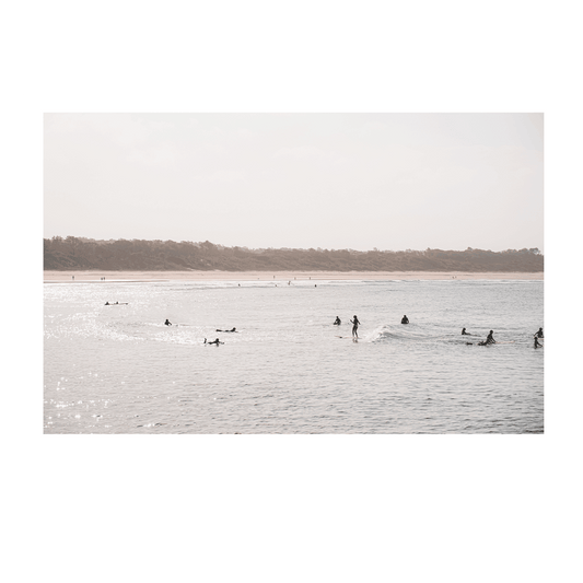 A serene sunset surf scene at Scotts Head, NSW, with surfers gliding across the shimmering ocean as golden light reflects off the water. The distant shoreline and soft pastel sky create a peaceful coastal moment.
