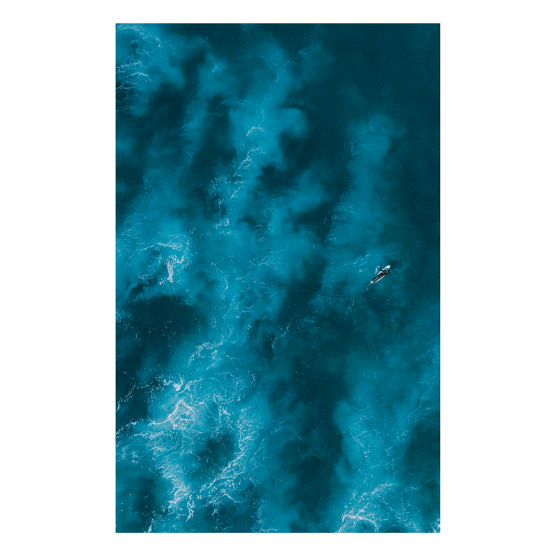 Experience the power of Angourie Beach with Brave – an aerial ocean print capturing a lone surfer paddling into vast, deep blue waters. A moody and dramatic seascape.