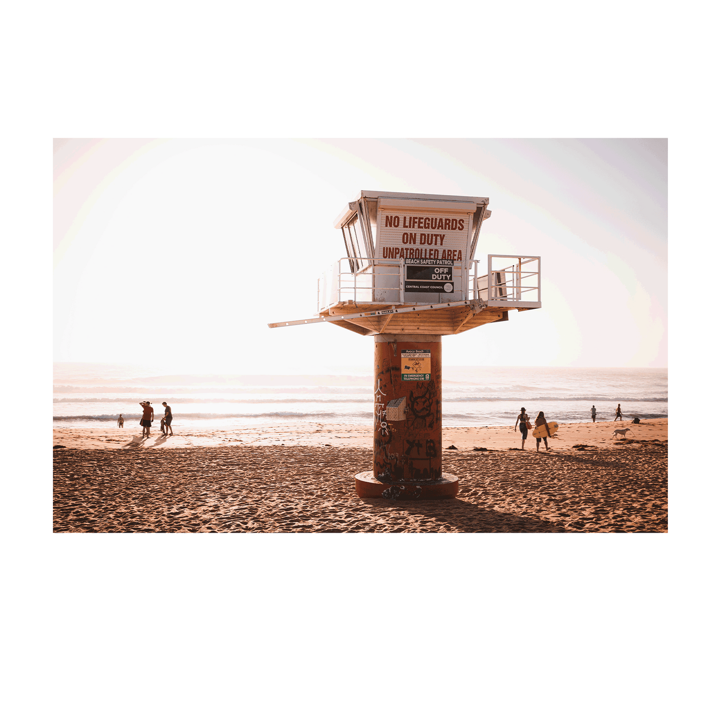 A lifeguard tower at Avoca Beach, NSW, with graffiti, warm sunlight, and beachgoers walking along the sandy shore.