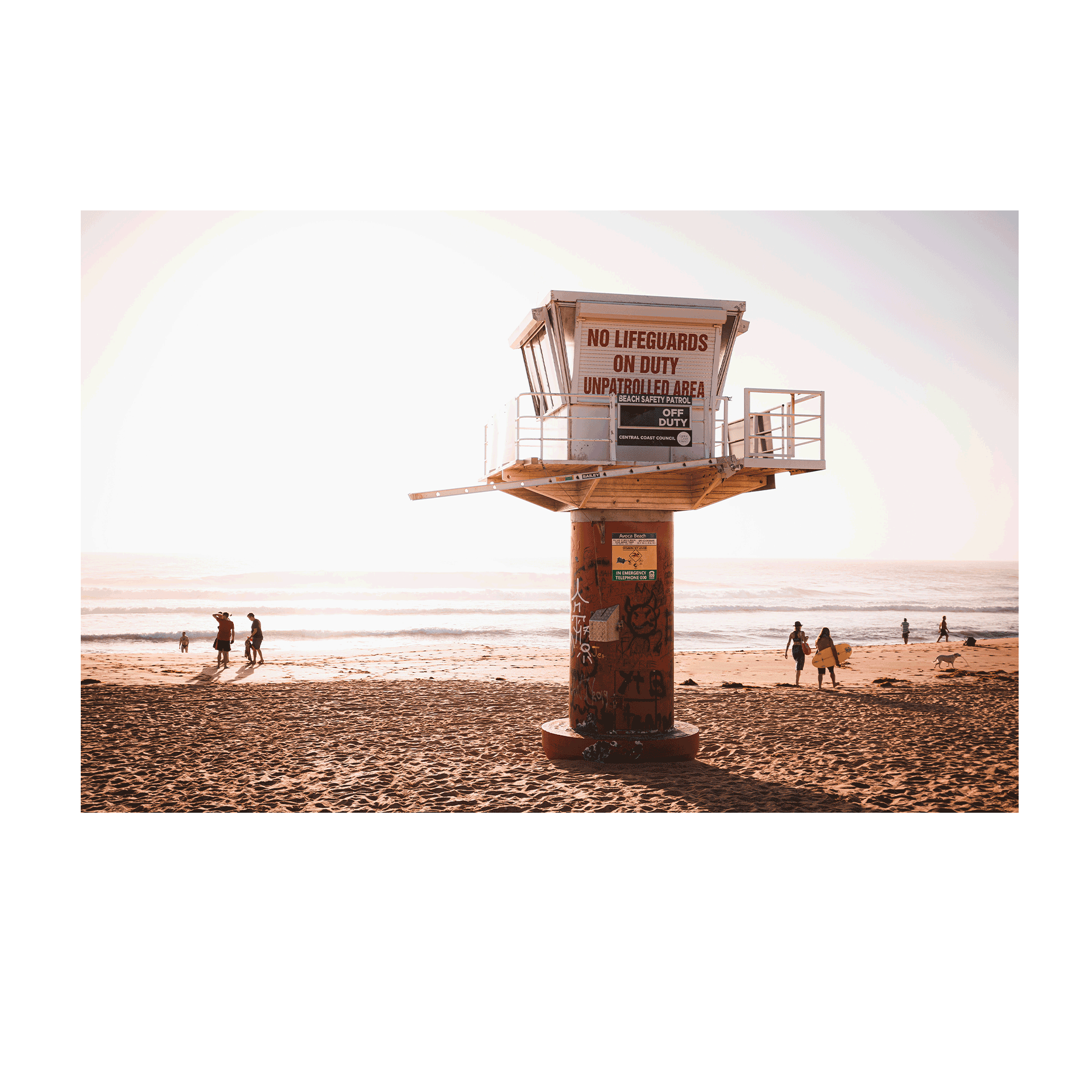 A lifeguard tower at Avoca Beach, NSW, with graffiti, warm sunlight, and beachgoers walking along the sandy shore.