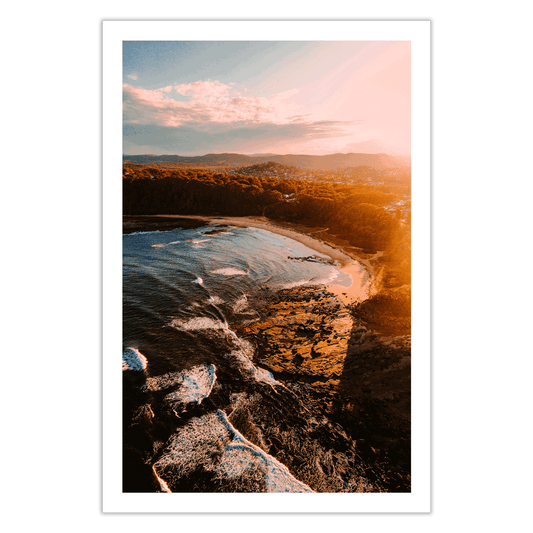 Aerial view of Bateau Bay, NSW, capturing golden light reflecting on the rugged coastline as waves crash against the shore at sunset.