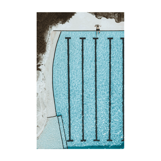 A striking aerial view of Bondi’s ocean pool, featuring a lone figure standing at the edge of crystal-clear turquoise waters with bold lane markings.