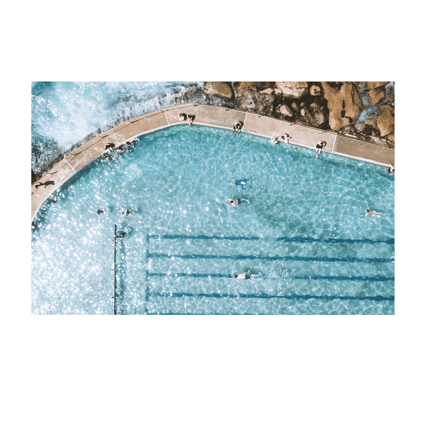 A sunlit aerial view of Bronte’s ocean pool in Sydney, featuring swimmers enjoying crystal-clear turquoise water with waves gently lapping at the pool’s edge.