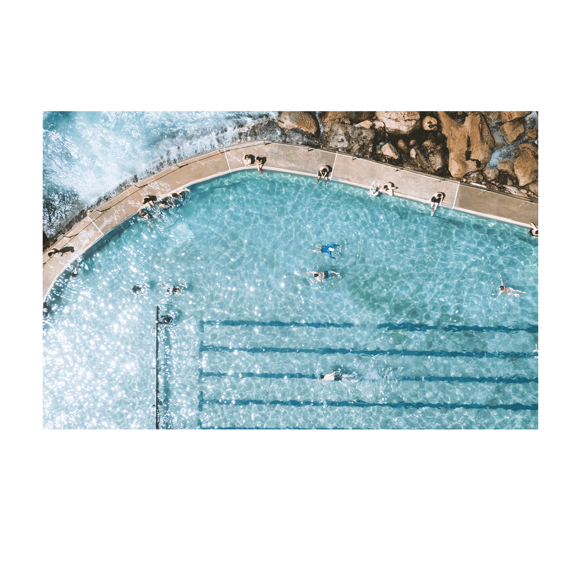 A sunlit aerial view of Bronte’s ocean pool in Sydney, featuring swimmers enjoying crystal-clear turquoise water with waves gently lapping at the pool’s edge.