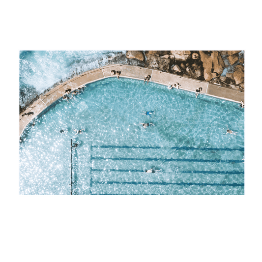 A sunlit aerial view of Bronte’s ocean pool in Sydney, featuring swimmers enjoying crystal-clear turquoise water with waves gently lapping at the pool’s edge.