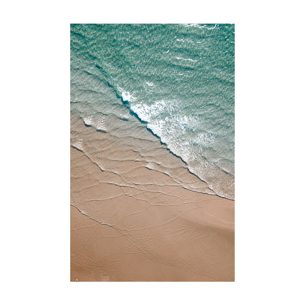 An aerial view of Byron Bay Beach, NSW, featuring turquoise waters gently meeting soft golden sands, with foam lapping at the shore.
