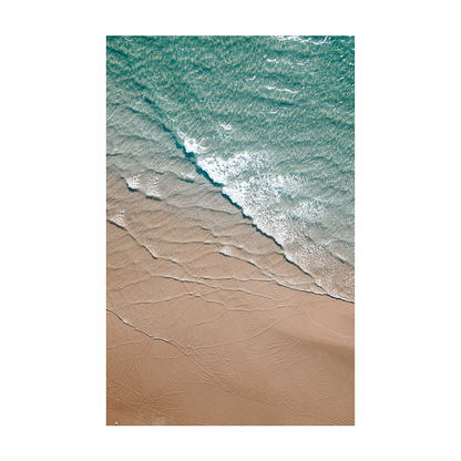 An aerial view of Byron Bay Beach, NSW, featuring turquoise waters gently meeting soft golden sands, with foam lapping at the shore.
