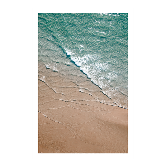 An aerial view of Byron Bay Beach, NSW, featuring turquoise waters gently meeting soft golden sands, with foam lapping at the shore.
