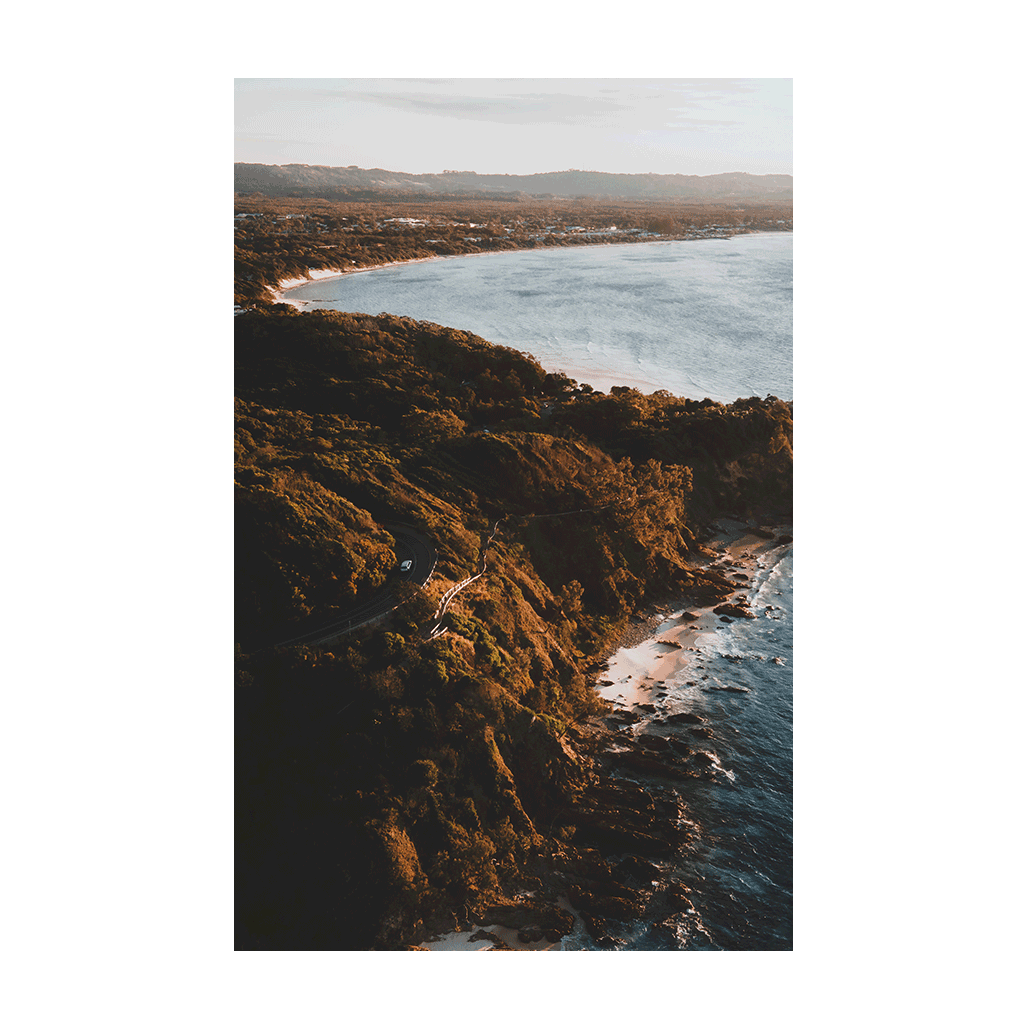 A stunning coastal print of Byron Bay, NSW, featuring a winding road along the cliffs at golden hour, with lush greenery and the ocean in the background.