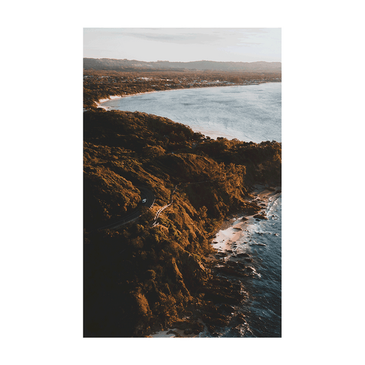 A stunning coastal print of Byron Bay, NSW, featuring a winding road along the cliffs at golden hour, with lush greenery and the ocean in the background.