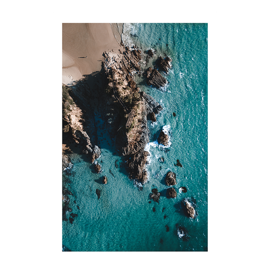 An aerial view of The Pass, Byron Bay, NSW, showcasing turquoise waters, rocky outcrops, and a lone figure walking along the beach.
