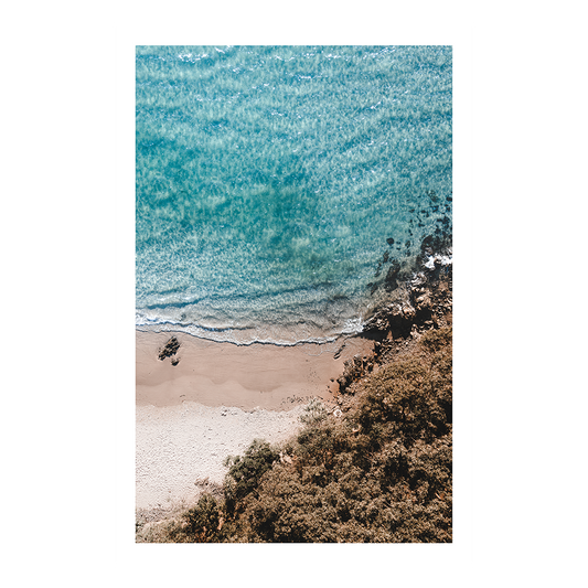 An aerial view of Byron Bay Beach, NSW, with turquoise waters meeting the sandy shore, surrounded by lush greenery and rocky outcrops.
