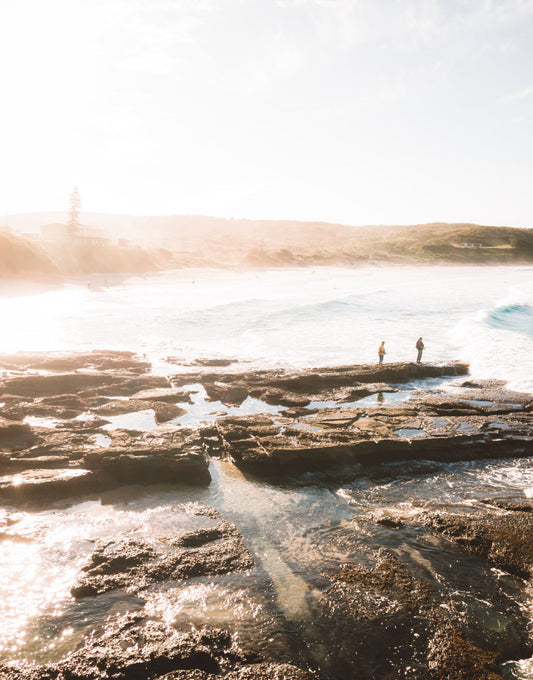 Aerial view of Catherine Hill Bay’s rocky shoreline with two fishermen standing on the rocks, surrounded by shimmering tidal pools and soft morning light.