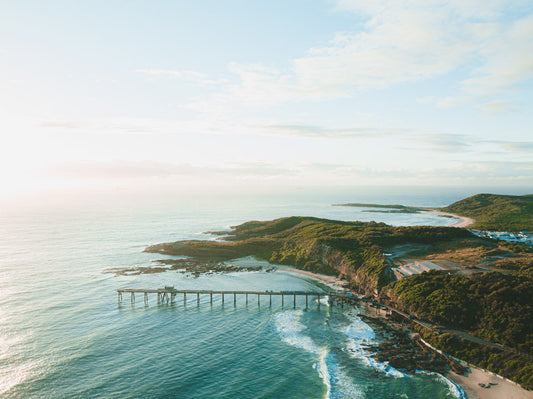 Aerial view of Catherine Hill Bay, NSW, showcasing the coastline with lush green hills, sparkling waters, and the iconic jetty under soft sunlight.
