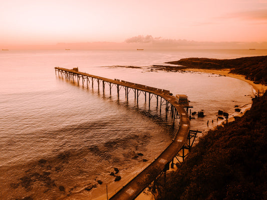 Aerial view of Catherine Hill Bay, NSW, showcasing the curved jetty extending over calm waters under a warm, golden sky.
