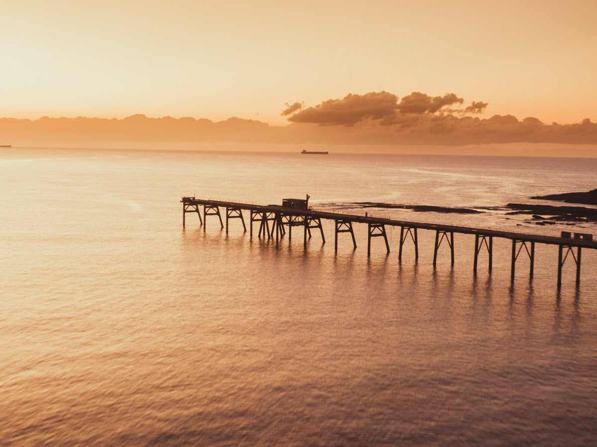 Aerial view of Catherine Hill Bay’s jetty at sunrise, with vibrant orange skies and golden reflections on tranquil waters.
