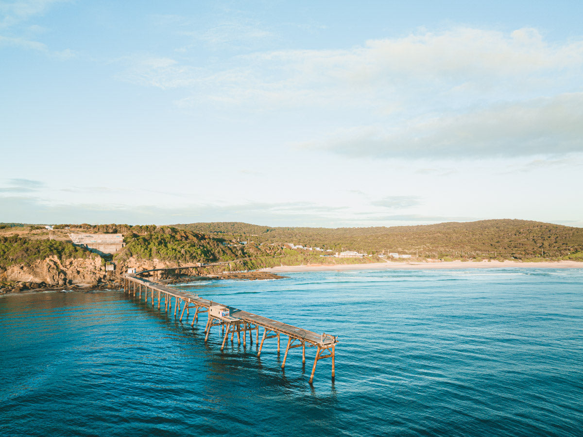 Aerial view of Catherine Hill Bay, NSW, showcasing the iconic jetty extending over vibrant blue waters with lush hills and sandy shores in the background.
