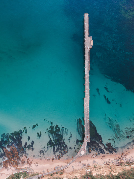 Aerial view of Catherine Hill Bay, NSW, showcasing the jetty extending over turquoise waters with a detailed view of the rocky shoreline and sandy beach.