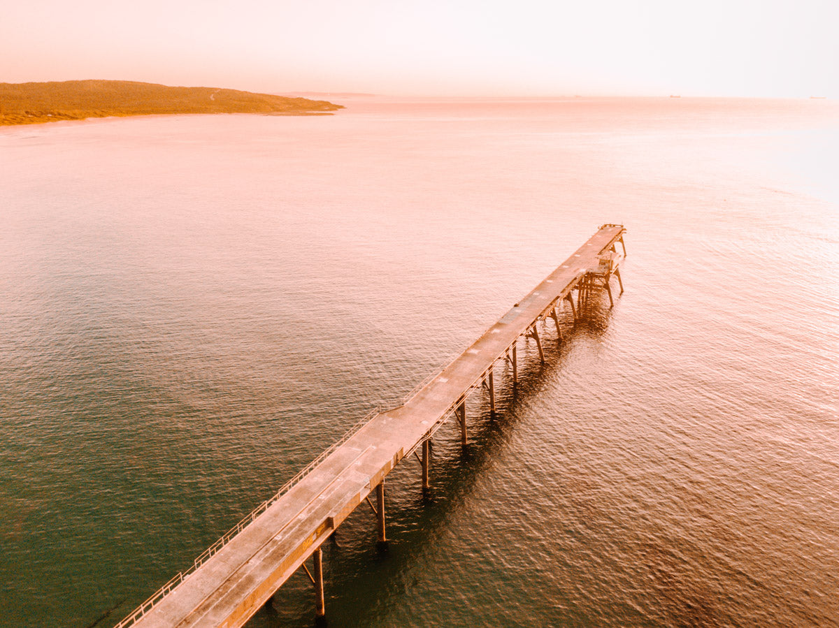 Aerial view of Catherine Hill Bay, NSW, featuring the jetty stretching into calm waters with warm, golden light reflecting off the ocean.
