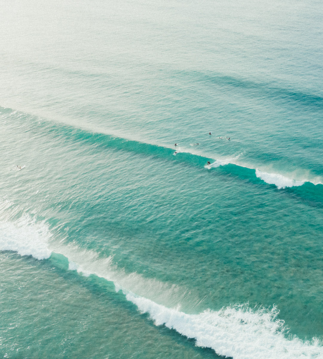 Aerial view of Catherine Hill Bay, NSW, showcasing surfers riding gentle turquoise waves under a soft and luminous sky.