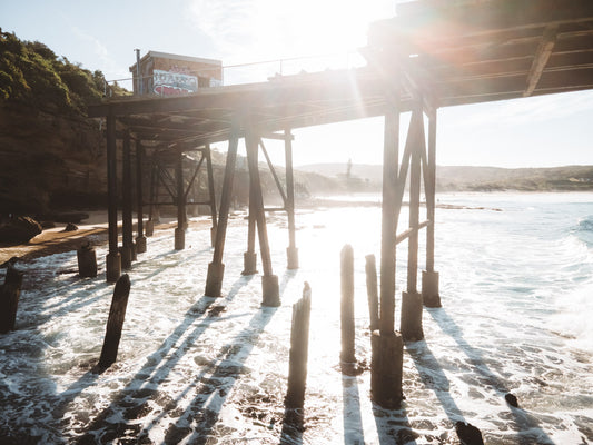 A unique view under Catherine Hill Bay’s jetty, showcasing weathered timber pylons, light and shadow patterns, and waves crashing against the shore.