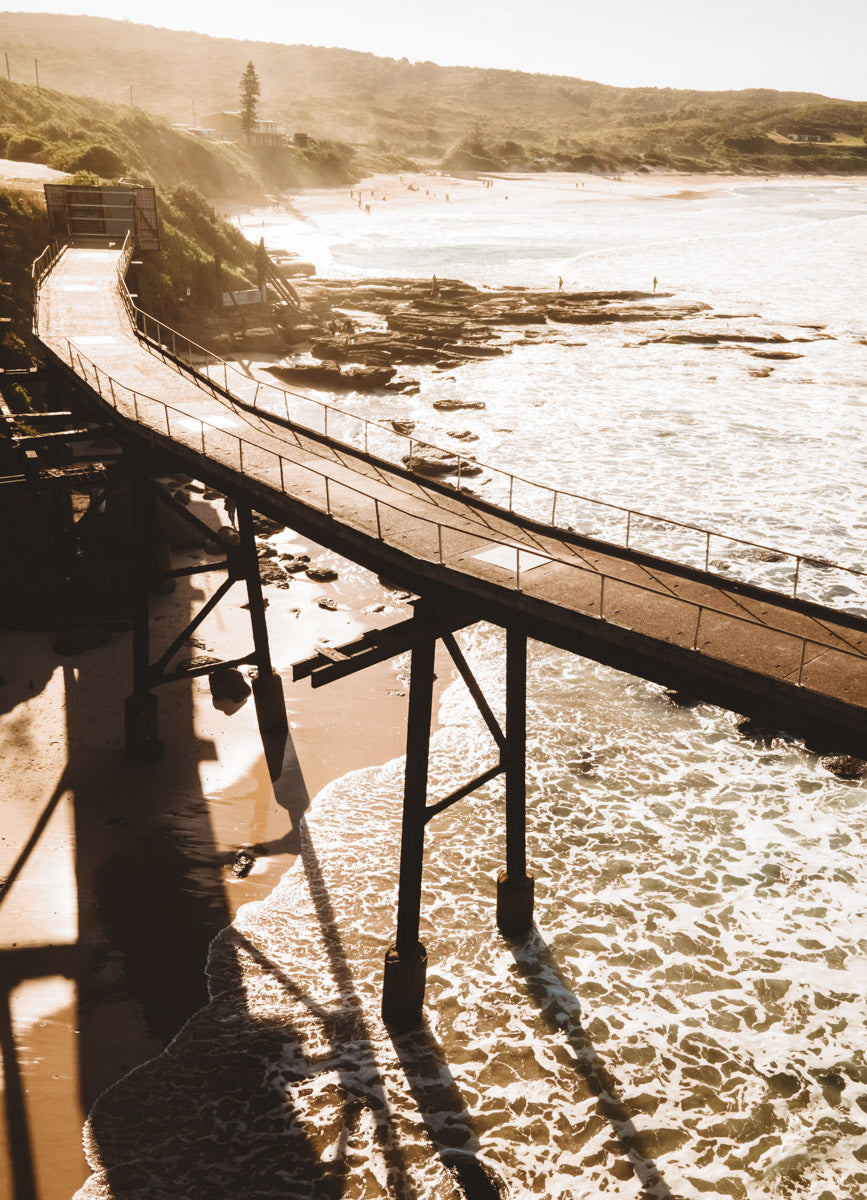 Close-up aerial view of Catherine Hill Bay’s historic jetty, with warm sunlight casting shadows over golden sands and the gentle waves below.
