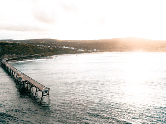 Aerial view of Catherine Hill Bay, NSW, featuring the iconic coal-loading jetty, calm waters, and surrounding green hills during a soft sunrise.
