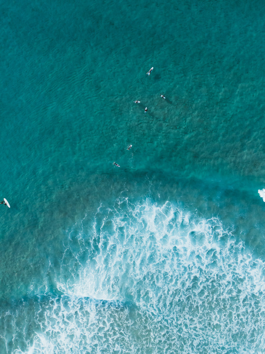 Aerial view of Catherine Hill Bay, NSW, featuring surfers in turquoise waters with white waves breaking and rippling across the ocean’s surface.