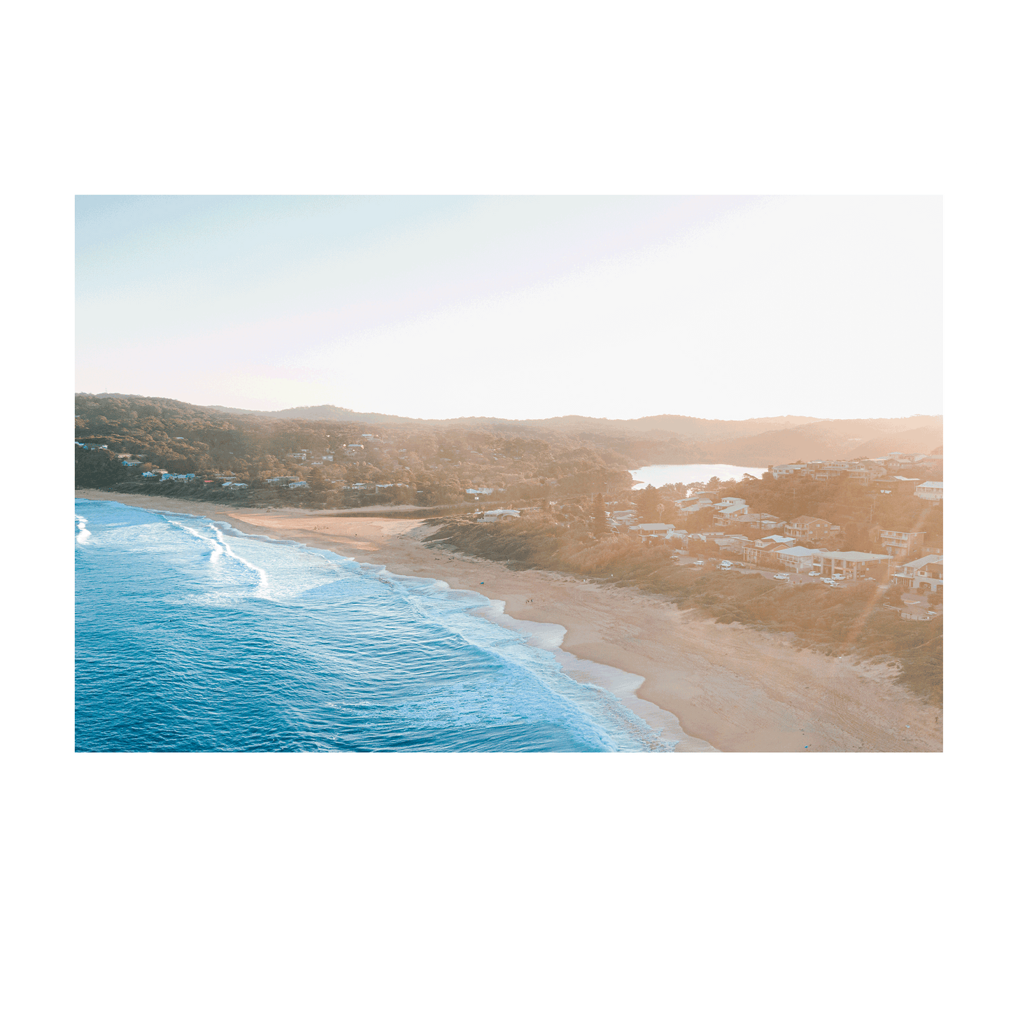 Aerial view of Copacabana Beach, NSW, at sunset, featuring rolling waves, golden sand, and a warm coastal glow over beachside homes.
