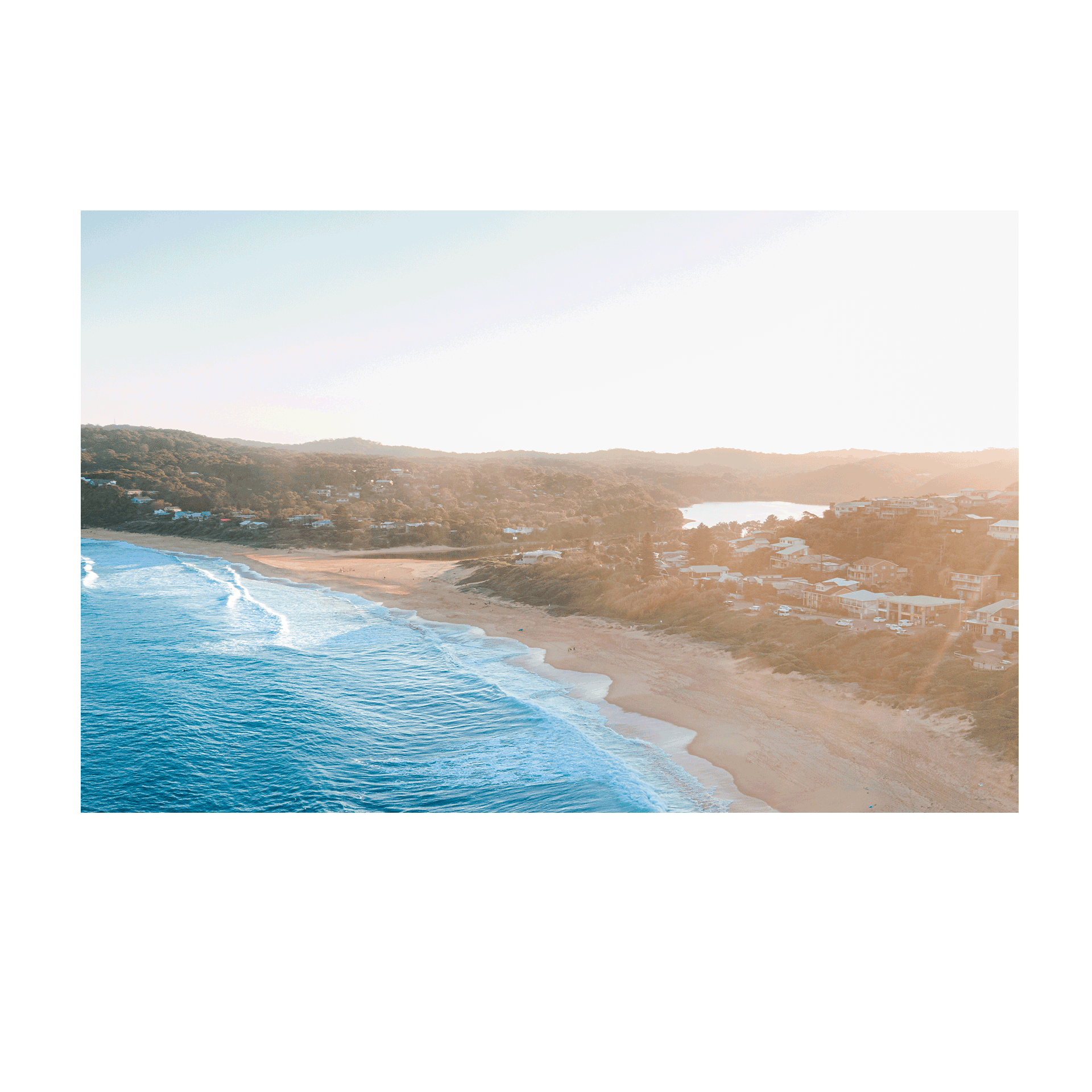 Aerial view of Copacabana Beach, NSW, at sunset, featuring rolling waves, golden sand, and a warm coastal glow over beachside homes.
