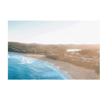 Aerial view of Copacabana Beach, NSW, at sunset, featuring rolling waves, golden sand, and a warm coastal glow over beachside homes.
