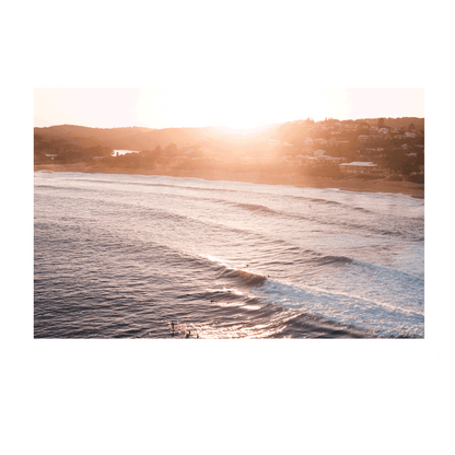 Aerial view of Copacabana Beach, NSW, at sunset, featuring rolling waves glowing under the setting sun with a warm coastal backdrop.
