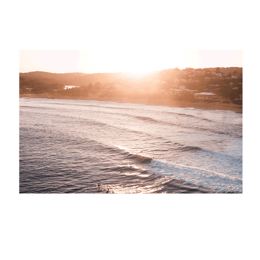 Aerial view of Copacabana Beach, NSW, at sunset, featuring rolling waves glowing under the setting sun with a warm coastal backdrop.
