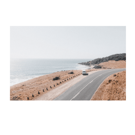 A scenic aerial view of a van driving along a coastal road at Racecourse, Crescent Head, NSW, framed by golden grasslands and ocean views.
