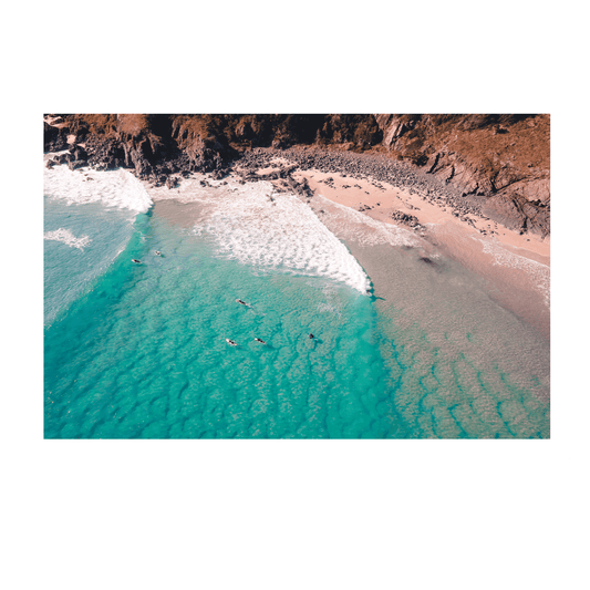 A stunning aerial view of Racecourse, Crescent Head, NSW, featuring surfers paddling in turquoise waves near a rugged coastline.
