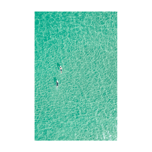 Aerial view of two surfers paddling through the crystal-clear turquoise waters of Forresters Beach, NSW.
